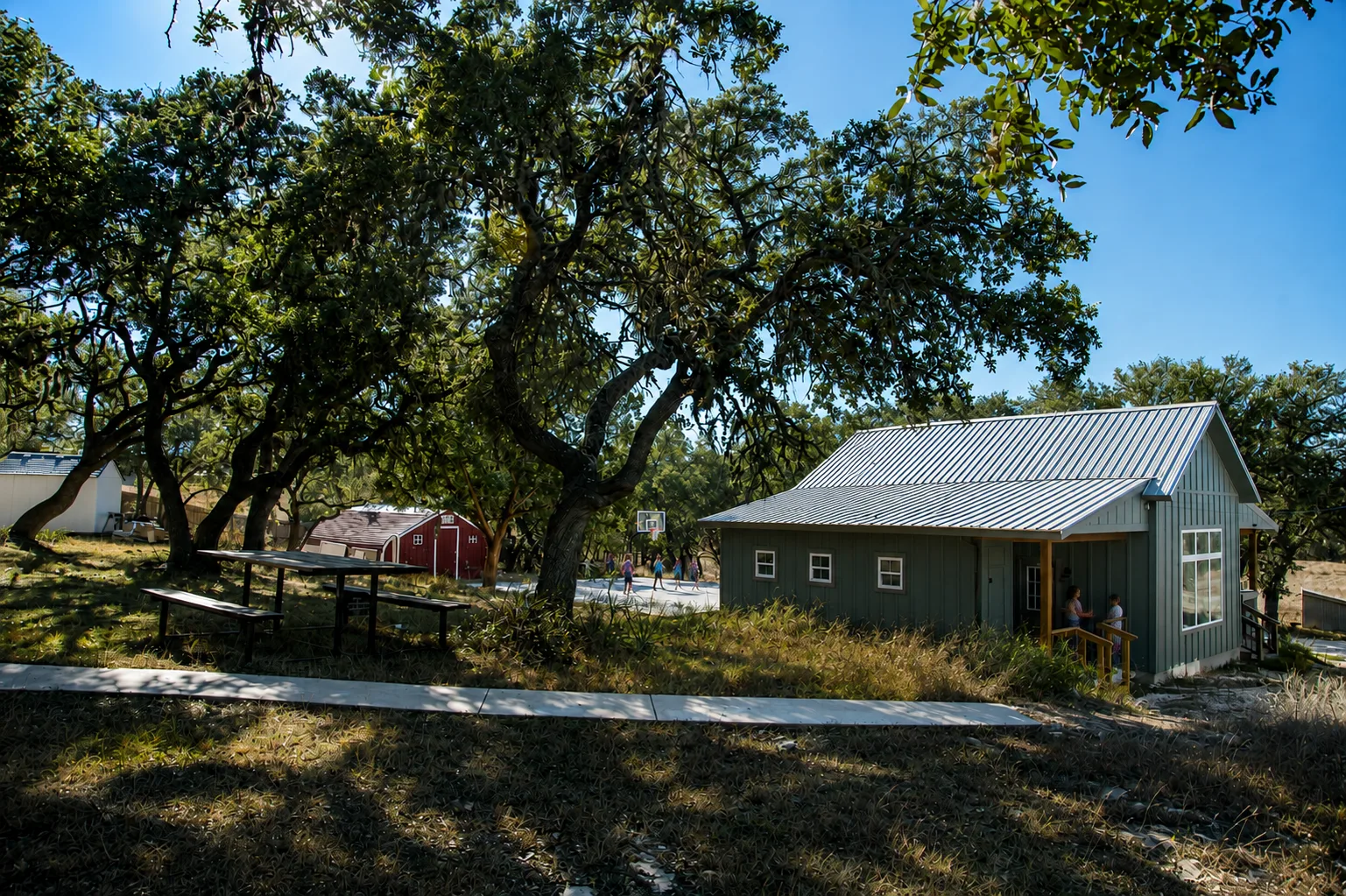 Green board-and-batten cottage on the Transformation House campus, shaded by live oak trees, with children playing basketball in the background.